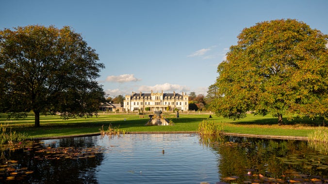 View of Dyffryn House from the fountain pool, two trees with autumnal-coloured leaves are either side of the fountain pool.